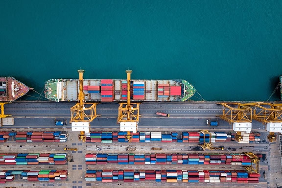 Aerial view of a cargo ship docked at a port with cranes loading and unloading shipping containers. Rows of containers are stacked along the docks.